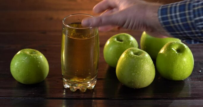 Man's hand takes the glass with apple juce from the table. There are fresh green apples