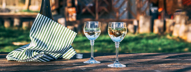 Two glasses of white wine and a bottle on an old wooden table in the courtyard of the house, a...