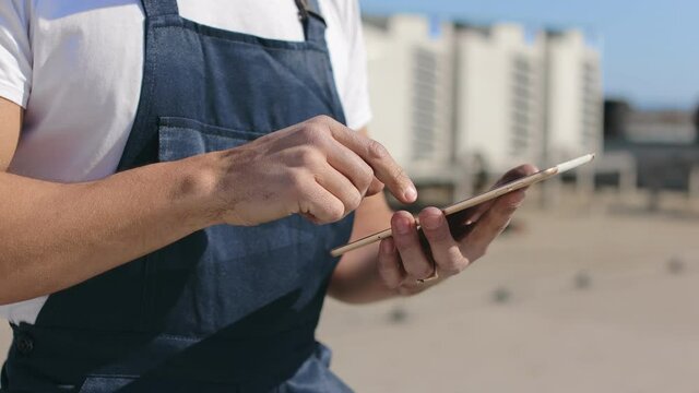 Close-up focus on hands of an engineer who is holding a tablet and working on it. He's on the roof of a business center. He is wearing work clothes. Air conditioning systems in the background. Nice