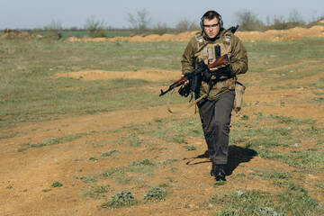 armed young white man in military uniform wearing headphones and glasses at the training ground
