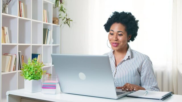 African young woman greets with her hand and talks while looking into the monitor screen. Webinar, conference remotely via video call. A black student is studying online at home. 4K.