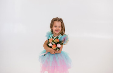cute little blonde girl in a tulle dress stands and holds a bouquet of fresh flowers on a white...