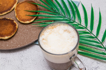 Tasty breakfast. A cup of coffee with milk or cream and marshmallows in a transparent mug with pancakes on a plate and with a palm branch. Nearby, coffee beans and marshmallows were scattered against 