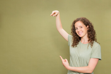 Portrait young redhead girl with curly hair smiling looking at camera pointing with both hands wearing green t-shirt on green background