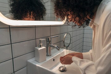 Young mixed race woman washing hands and face in a modern washroom. Curly hair female getting ready...