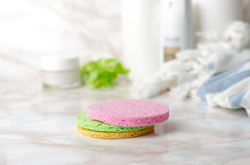 Closeup of stack of colorful scrub sponges for face, towel, bottles of cosmetic products on the bright surface