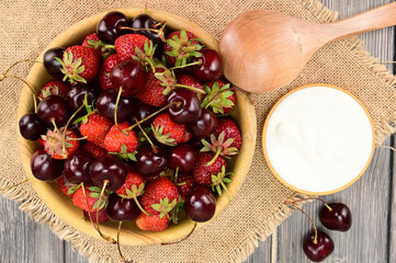 Wooden plate with strawberries and cherries on burlap on wooden boards. Nearby there is a saucer of sour cream and a wooden spoon