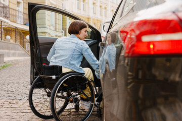 Brunette woman in wheelchair getting into car on city street - obrazy, fototapety, plakaty