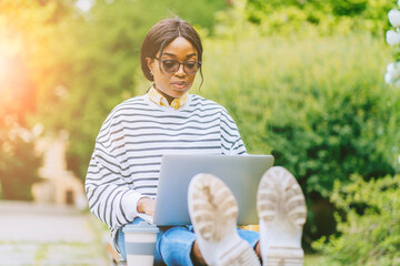 Pleased african american woman student in eyeglasses sitting on bench and using laptop preparing for exams in city park over flowering bush on background, outdoors.