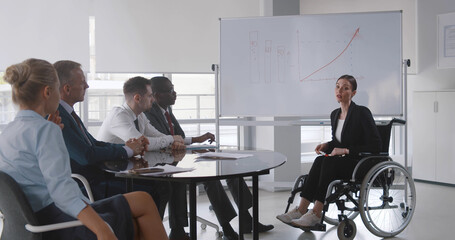 Young woman in wheelchair teaching group of business people at whiteboard in office