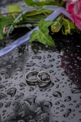 Wedding rings on the hood of the vehicle along with raindrops and part of the wedding bouquet.