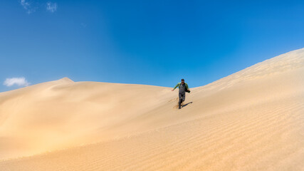 Fototapeta na wymiar A man walks down the dunes of the desert. The guy runs on the sand. Sunny day blue sky.