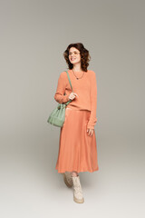 full length of stylish woman in glasses, sweater and silk skirt holding leather strap of bag on grey