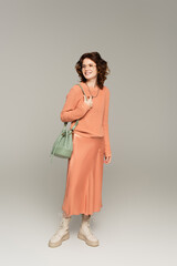 full length of happy and stylish woman in sweater and satin skirt holding leather strap of bag and standing on grey