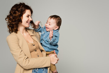cheerful toddler boy holding glasses while happy mother smiling isolated on grey