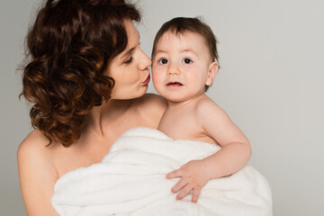 caring mother with bare shoulders kissing baby boy wrapped in towel isolated on grey