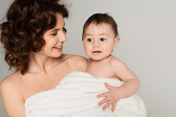 smiling mother with bare shoulders holding toddler son wrapped in towel isolated on grey