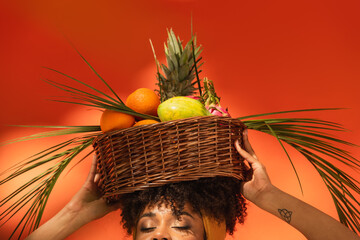 partial view of young african american woman with closed eyes holding basket with exotic fruits on head on orange