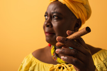 middle aged african american woman in yellow turban and blouse holding cigar on yellow