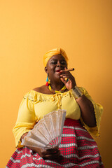 Obraz premium middle aged african american woman in bright clothes sitting with fan and smoking cigar on yellow