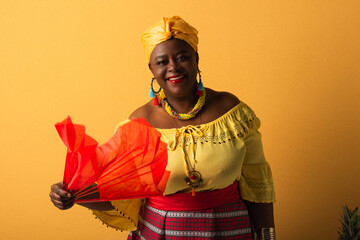 smiling middle aged african american woman in yellow turban and blouse holding red fan on yellow