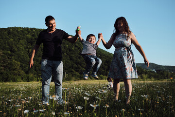 Mom, dad and son on walk in chamomile field in summer. Young parents hold their young son with both hands and swing him back and forth and the child laughs merrily with his mouth wide open.