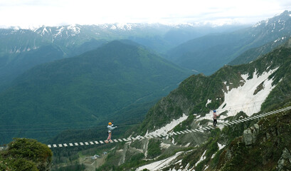 Tourist walks on rope suspension bridge on Rosa Peak (2320 m.) in Sochi, Russia. People on hanging footbridge above chasm, extreme attraction. Beautiful landscape, mountain peak in clouds and snow