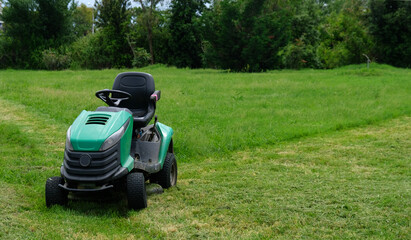 Fototapeta premium Green grass trimming with lawn mower. Tractor mowing grass in a public park. A dirty lawn tractor after mowing the lawn stands on the meadow. Professional lawn mower cuts the grass.
