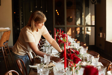 Caucasian girl florist decorator decorates the wedding table with red roses and eucalyptus in red boxes with red candles and chili and serves the table with glasses, plates and napkins