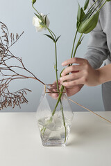 Cropped shot of a woman in with flowers and geometric glass vase with ice texture and golden rim. The lady in gray clothes is making floral arrangement of white flower and dried brown twig. 