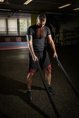 Fototapeta na wymiar Strong young bearded man working out with battle ropes a gym. Determined guy using battle rope while doing physical training. Hard workout