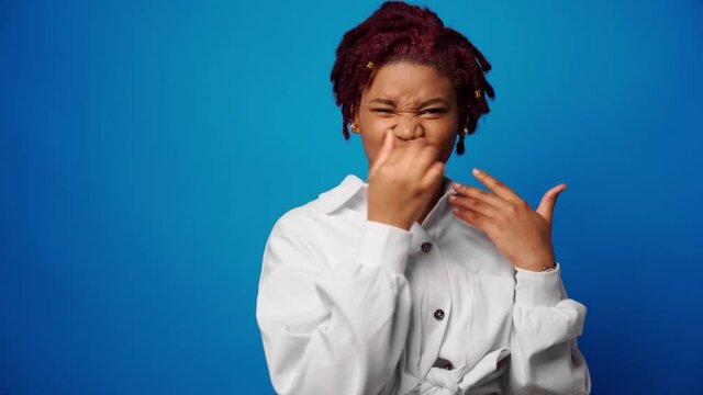 Afro woman in white shirt close nose because of a bad stinky smell, blue background