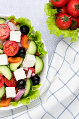 Greek salad or horiatiki salad made with pieces of tomatoes, cucumbers, onion, feta cheese, bell pepper slices, olives and dressed with olive oil. Vegetables on a white plate on a gray background.