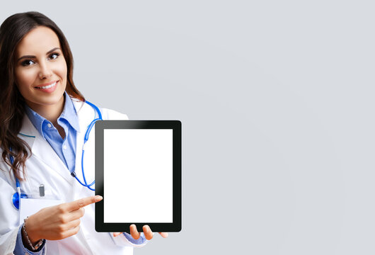 Portrait Image Of Happy Smiling Female Doctor Showing Pointing Tablet Pc With Copy Space Area For Some Text, Over Grey Colour Background. Medical Contact Center Concept Picture.