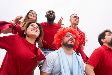 A group of fans of a soccer team celebrate a goal in the stands wearing red shirts while a fan of the opposing team mournfully laments.