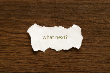 The question What's next is written on a white piece of paper on a dark table.