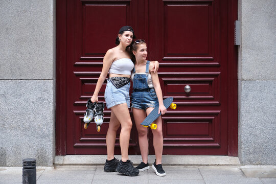 Two women holding a skateboard and inline skates in a city. Friends, urban life.