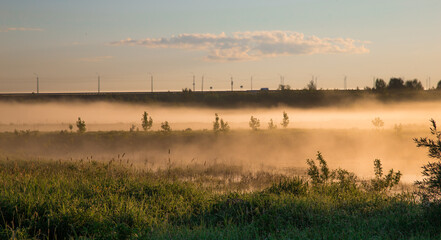 thick fog rises over fields in the rays of the rising sun on a warm summer morning