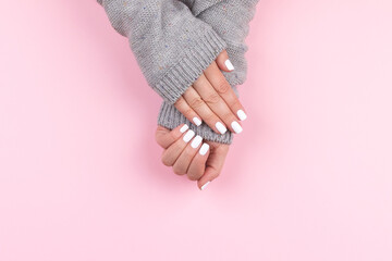 Female hands in gray knitted sweater with beautiful manicure - white nails on pink background,...