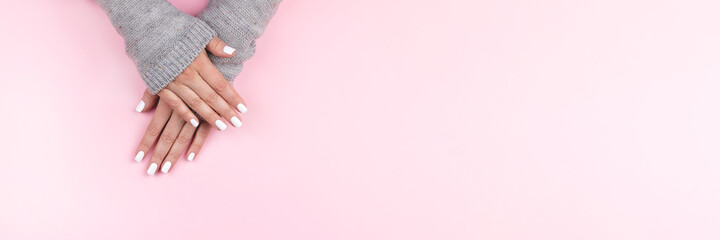 Female hands in gray knitted sweater with beautiful manicure - white nails on pink background, wide banner with copy space. Nail care concept