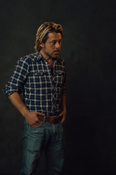 Man with blond hair in lumberjack shirt and jeans standing in front of a dark wall.