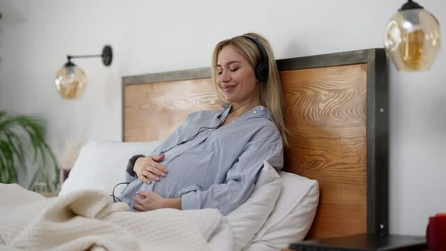 Happy smiling pregnant woman enjoying music in headphones lying on bed as unborn child pushing belly. Portrait of excited relaxed Caucasian blond beautiful woman enjoying pregnancy at home