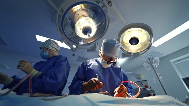 Medical team perform an operation under surgical lamp. Front view of a surgeon and female assistant doing a surgery using special medical tools.