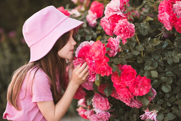 Cute child girl 5-6 year old wear hat and summer clothes posing with garden roses outdoors. Summer season. Childhood. Stylish toddler over nature background.