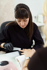 Woman manicurist wearing black gloves making manicure in luxury beauty salon with modern equipment, soft colors, nail service, perfect client hands.