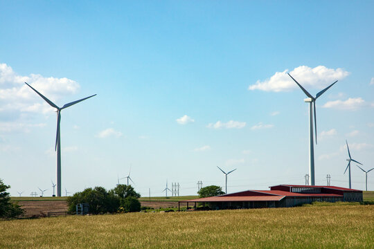 Wind Turbine Farm In An Expansive Green Field With Blue Sky With Puffy Clouds And The Roof Of A Grond Contact House