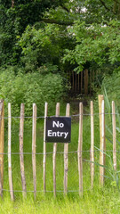 Rustic wooden chestnut paling fence blocking a rural public footpath. Generic black and white No Entry sign. Overgrown vegetation with trees. Portrait image with space for text. England. - 440307417