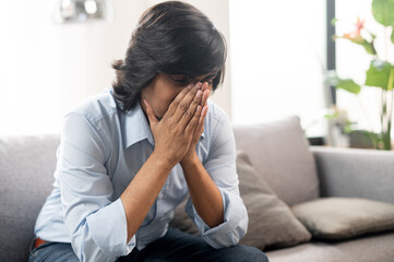 Unhappy young indian man sitting on the sofa, covered face with hands and feel frustration, tired and depressed eastern man alone at home, suffering from migraine
