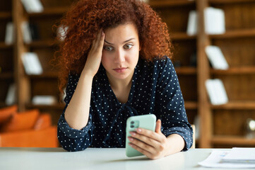 Worried red-haired young female freelancer or student with curly hair sitting at the desk, holding...