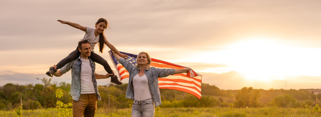 Young parents with their daughter holding American flag in countryside at sunset. Independence Day celebration